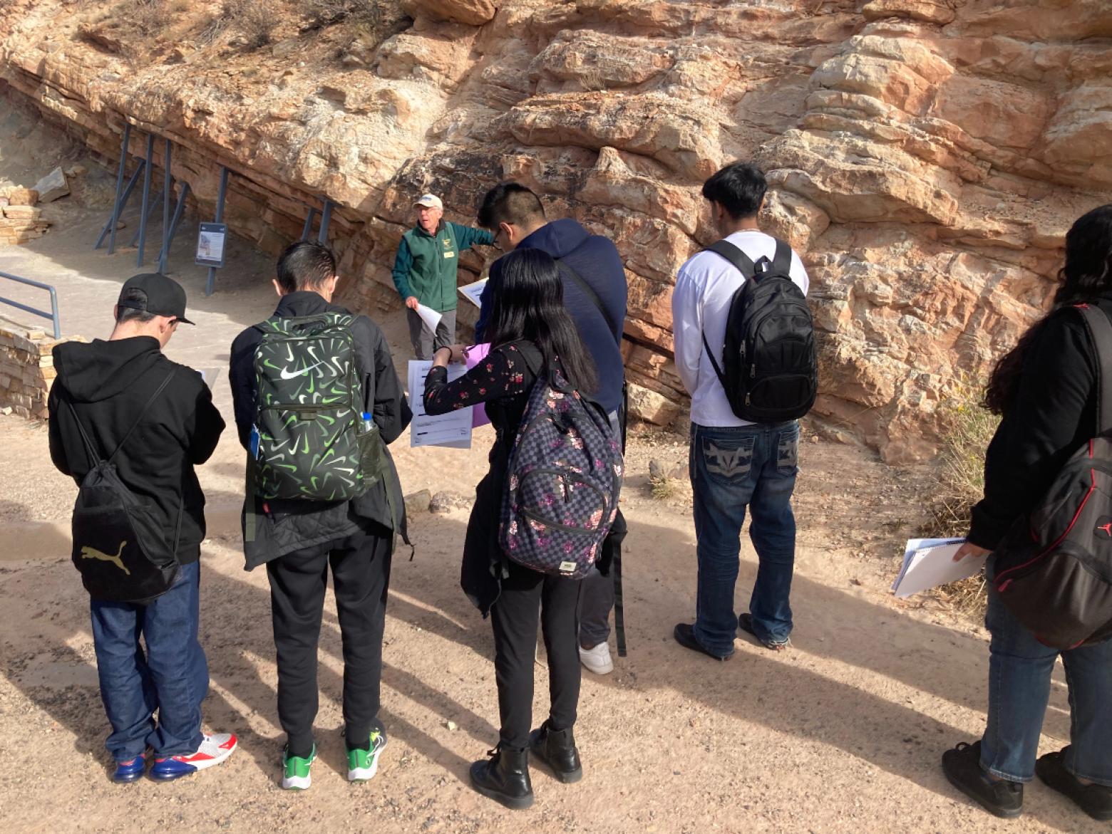 A group of students in backpacks stands listening in front of a rock formation
