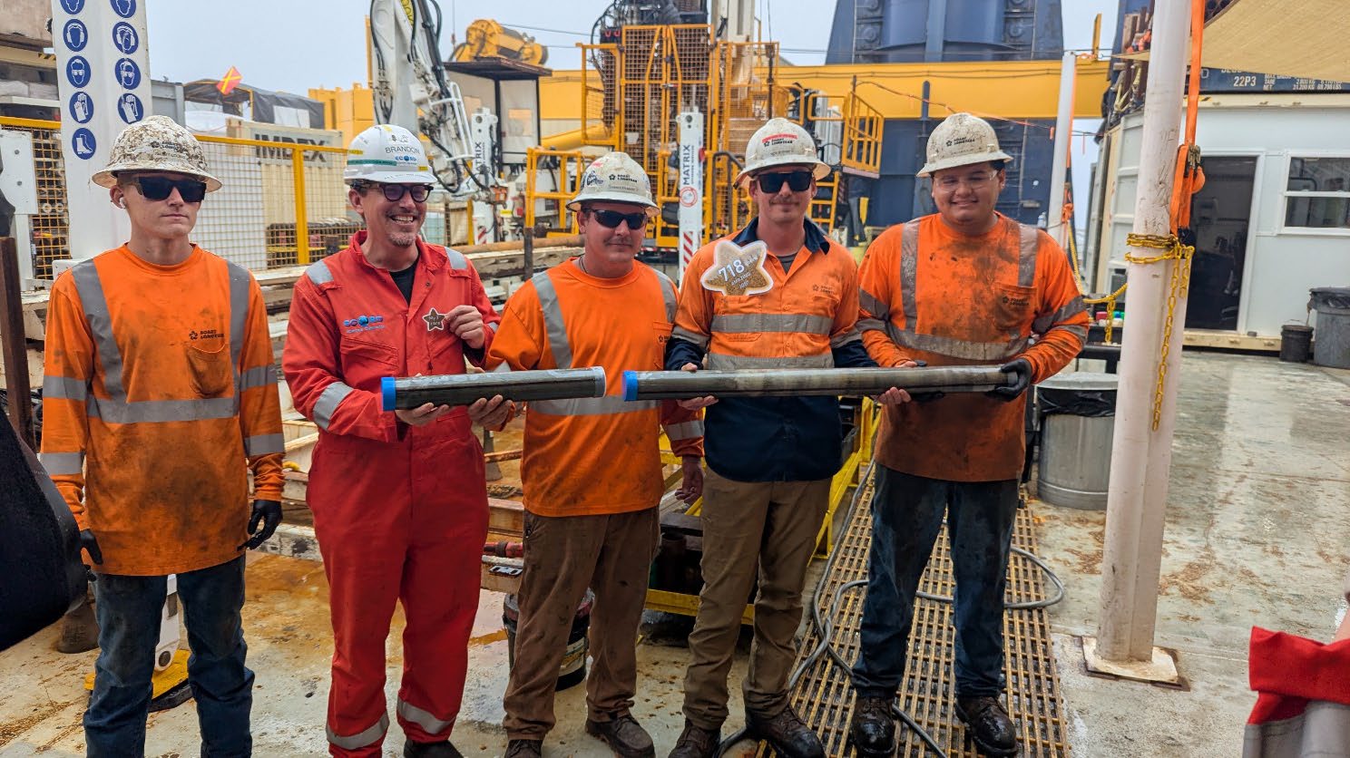 five men in safety orange hold metal tube holding final core from IODP³-NSF Expedition 501