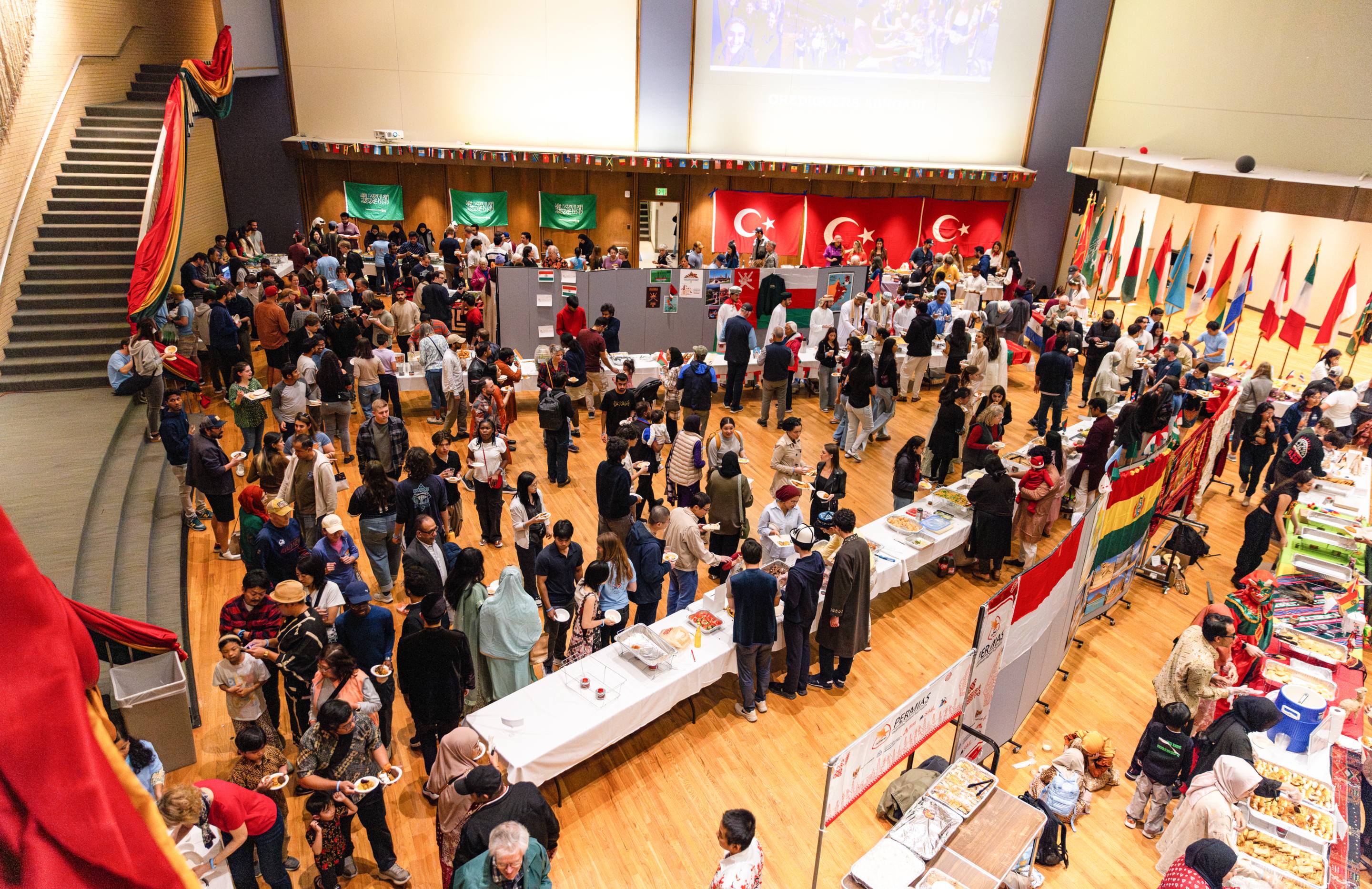 Elevated view of a large crowd gathered in an auditorium at Colorado School of Mines' International Day, with international flags hanging from the ceiling and lining the walls as students sample food at rows of tables.