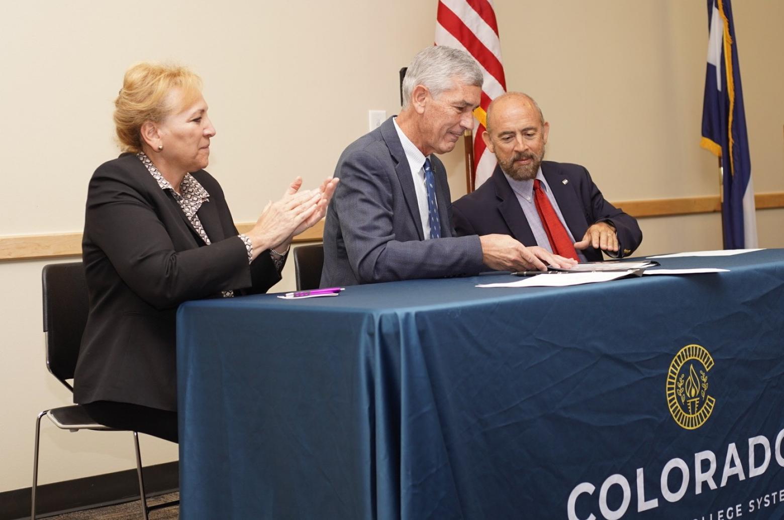 Angie Paccione, Paul Johnson and Joe Garcia sit at a table with the Community Colleges of Colorado logo on it