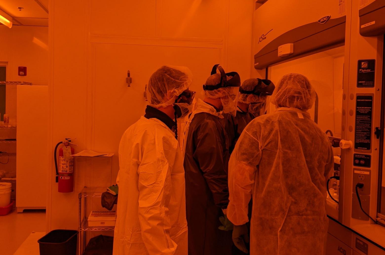 Four people in protective gear work in a cleanroom illuminated by orange light