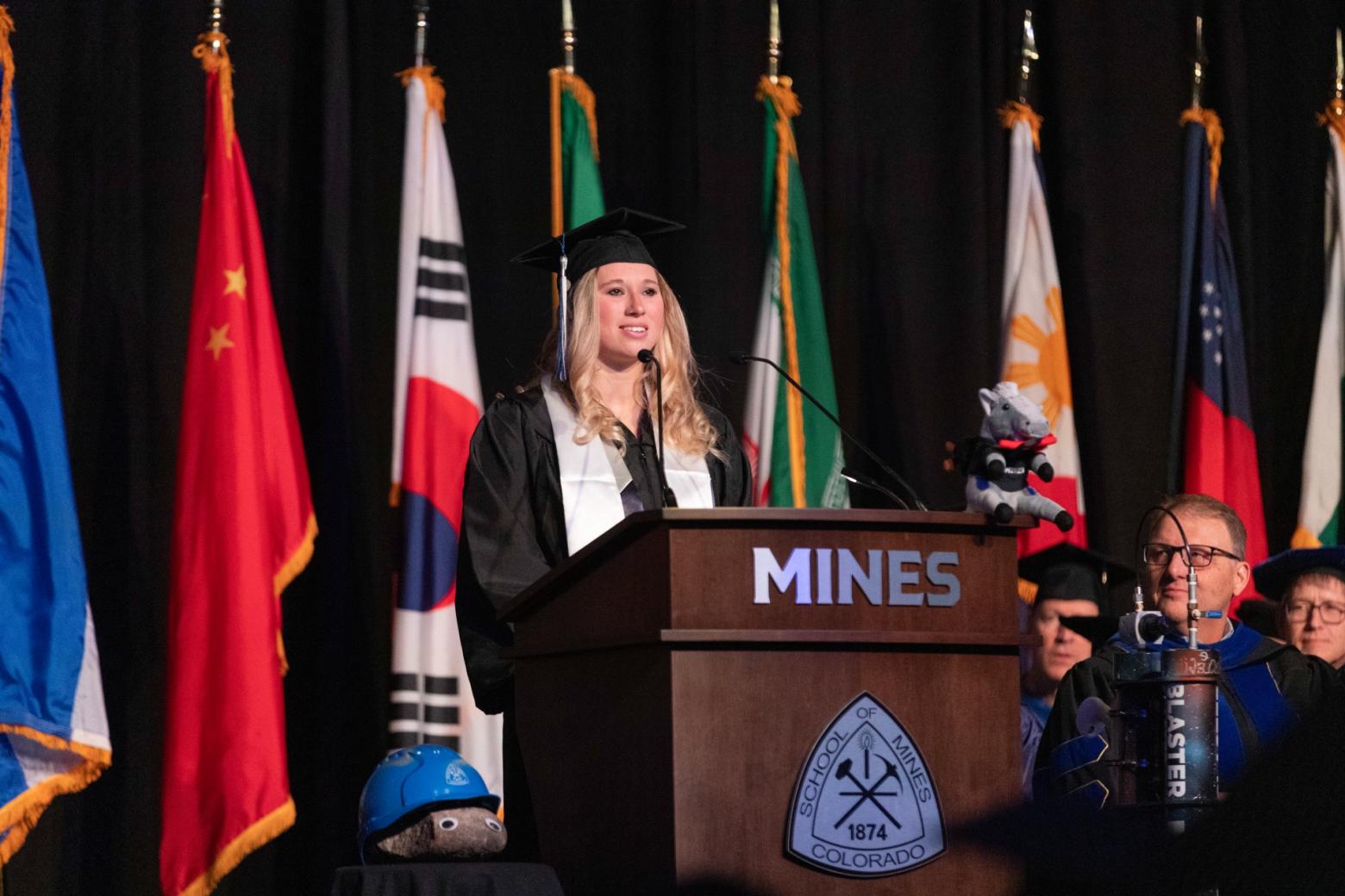 2023 Mines undergraduate speaker addresses the audience in her cap and gown at a podium.