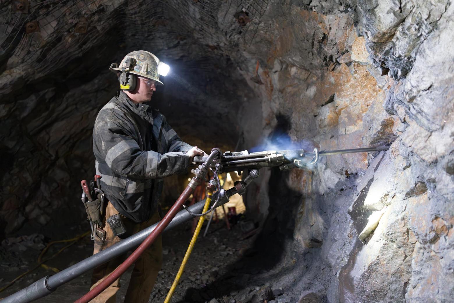 Miner wearing a helmet with light uses drilling equipment inside an underground rocky tunnel.