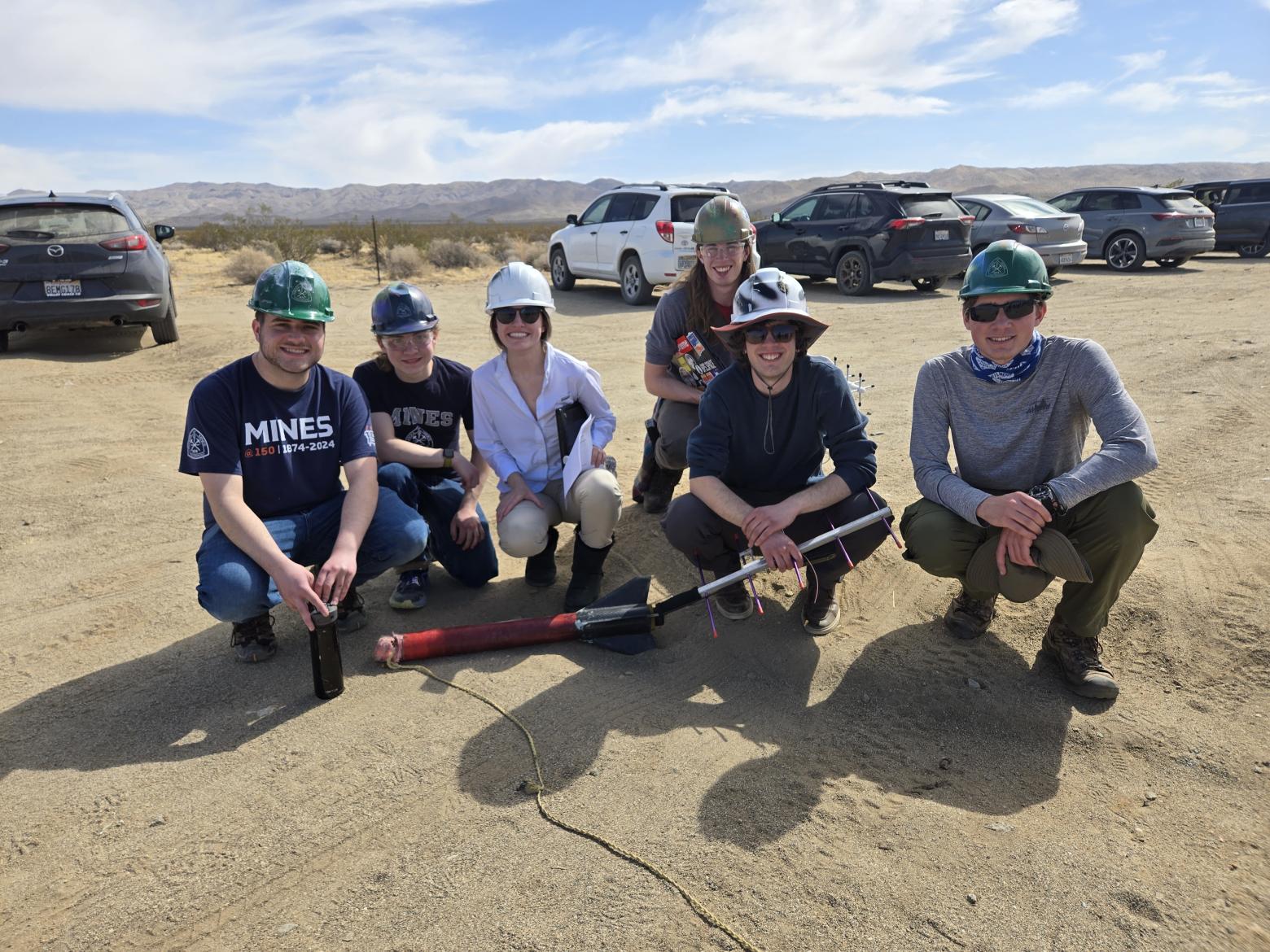 group of students in hard hats pose with rocket in gravel lot