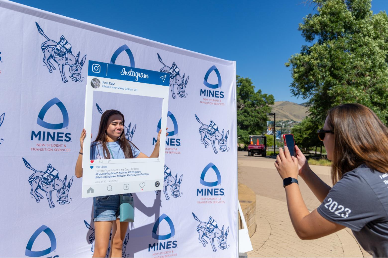 A Mines student holds up a large photo frame and stands in front of a backdrop with Mines logos to get her picture taken on the first day of class.