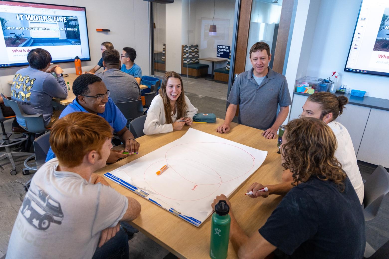 Group of students collaborating around a table with a large sheet of paper, whiteboards, and monitors in a modern meeting room.