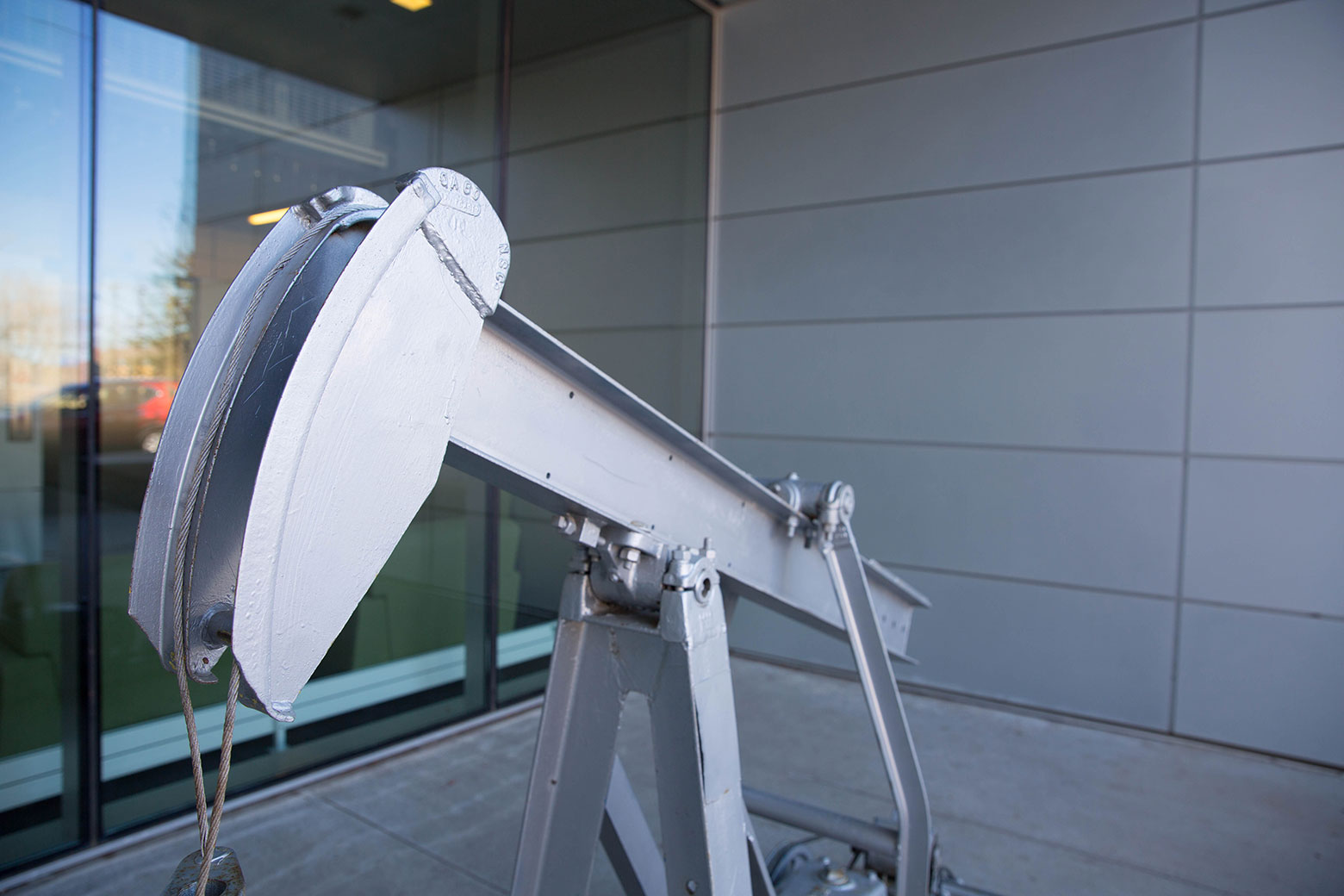 Small silver oil pump jack model displayed outdoors by a glass window and modern gray wall.