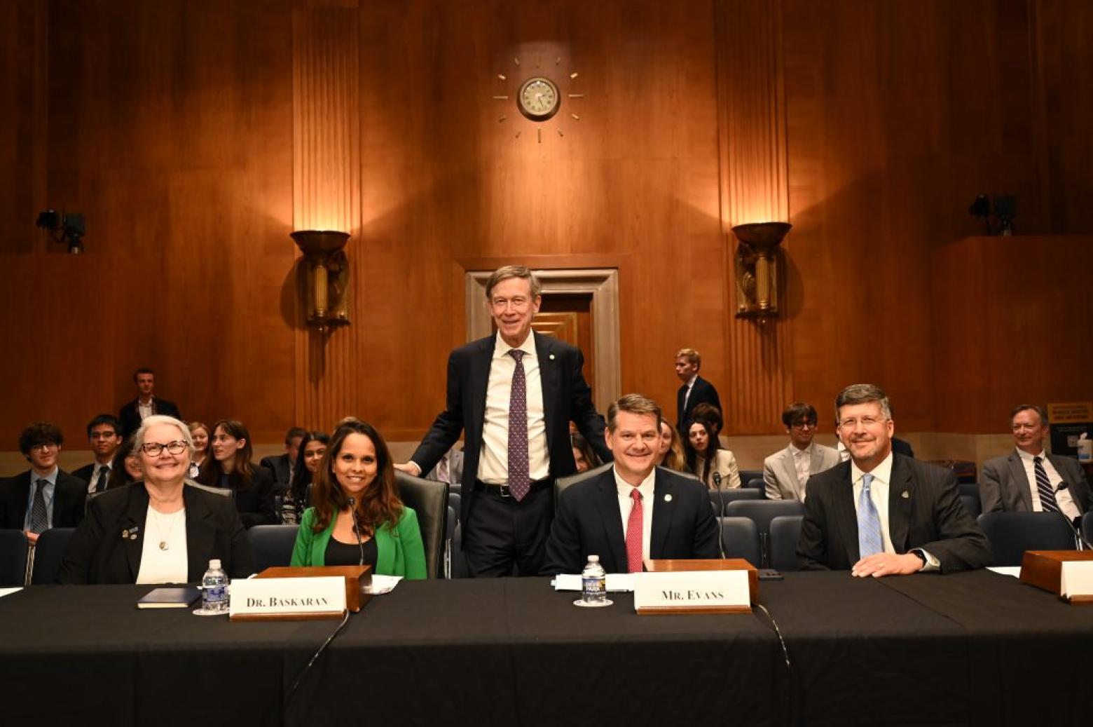 Bill Zisch sits at a table of experts with John Hickenlooper standing behind them