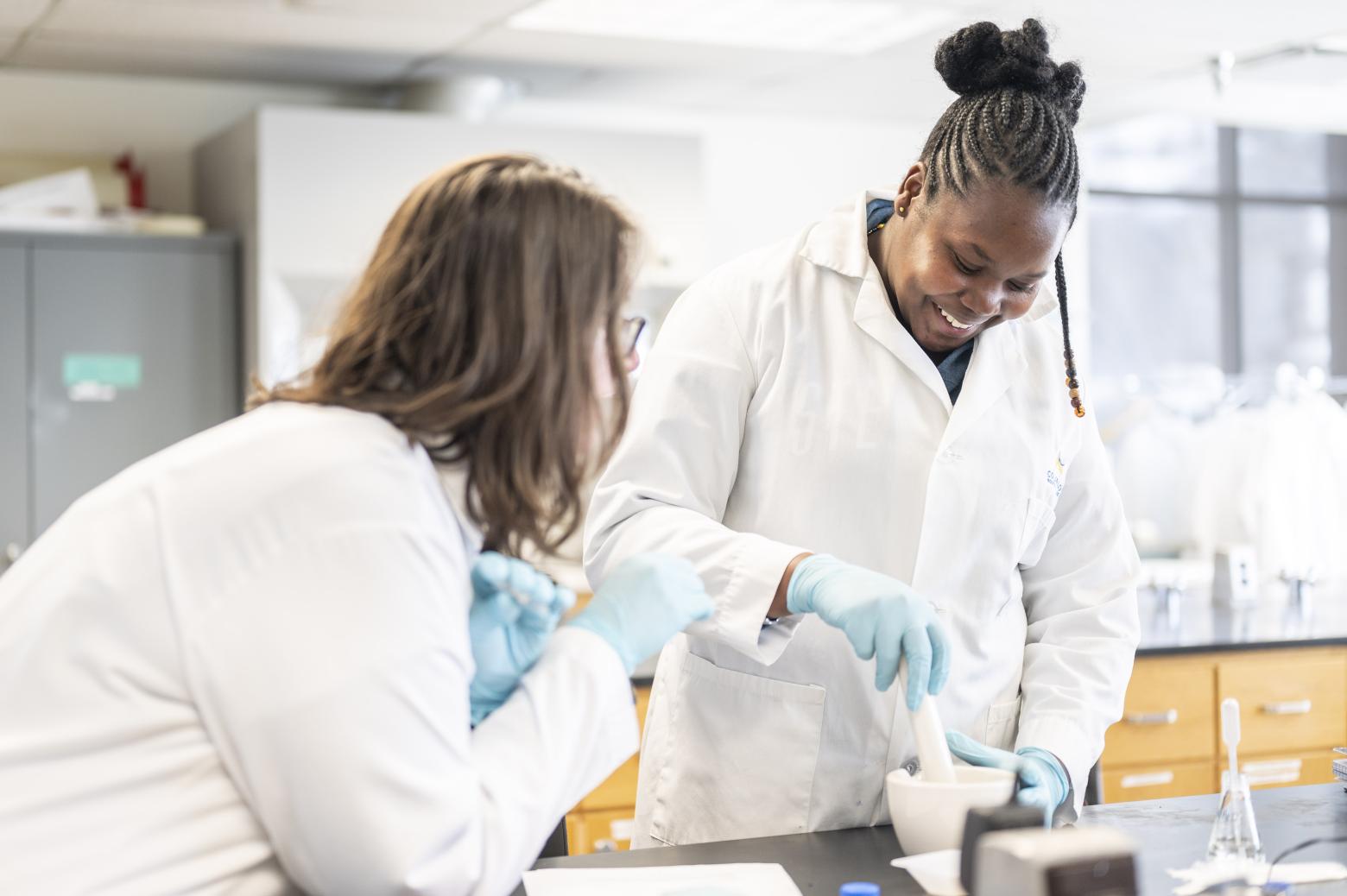 Two students in lab coats and gloves working with a mortar and pestle in a bright, organized laboratory.