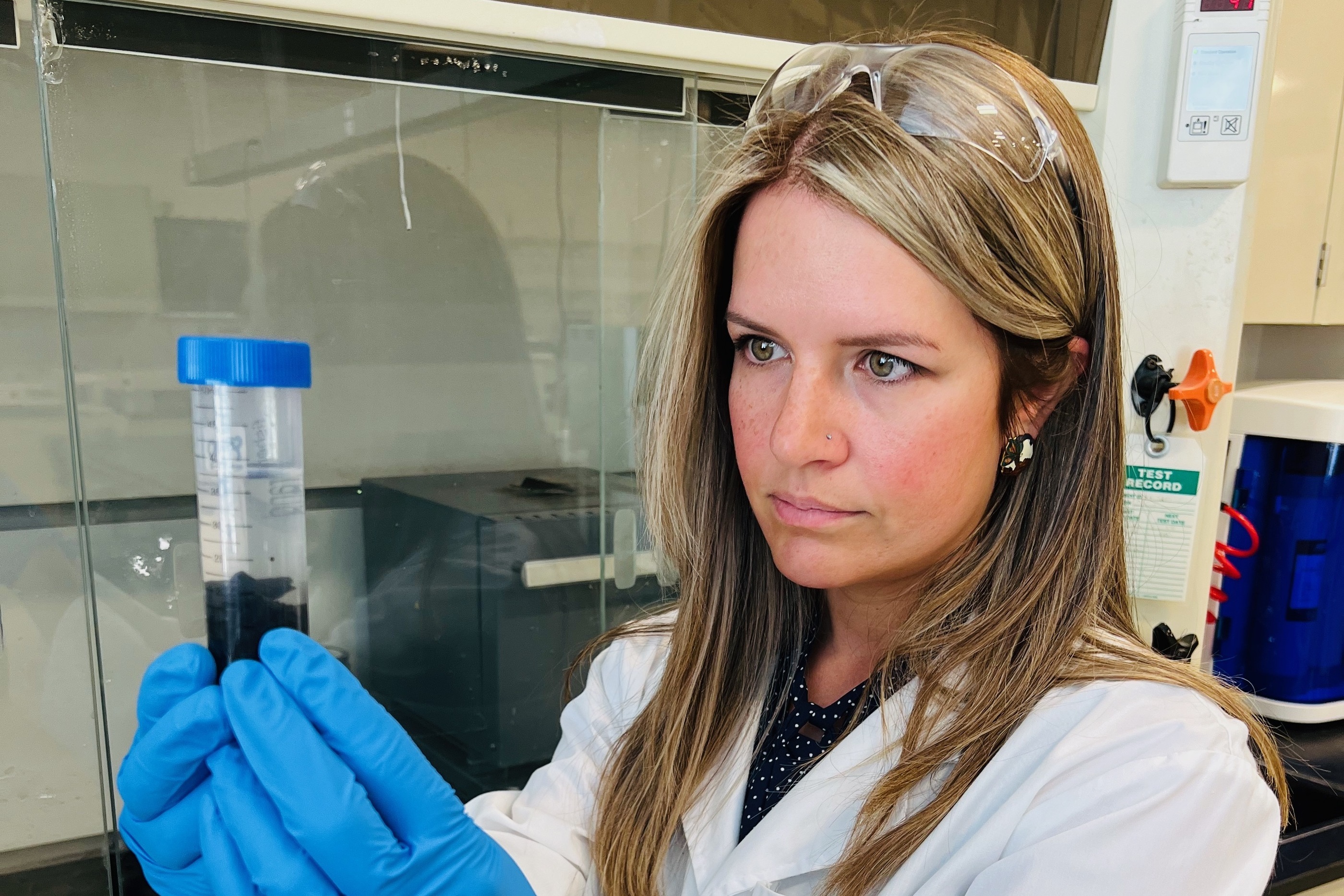 Lori Tunstall wears a white lab coat and blue gloves and looks at a vial of black material
