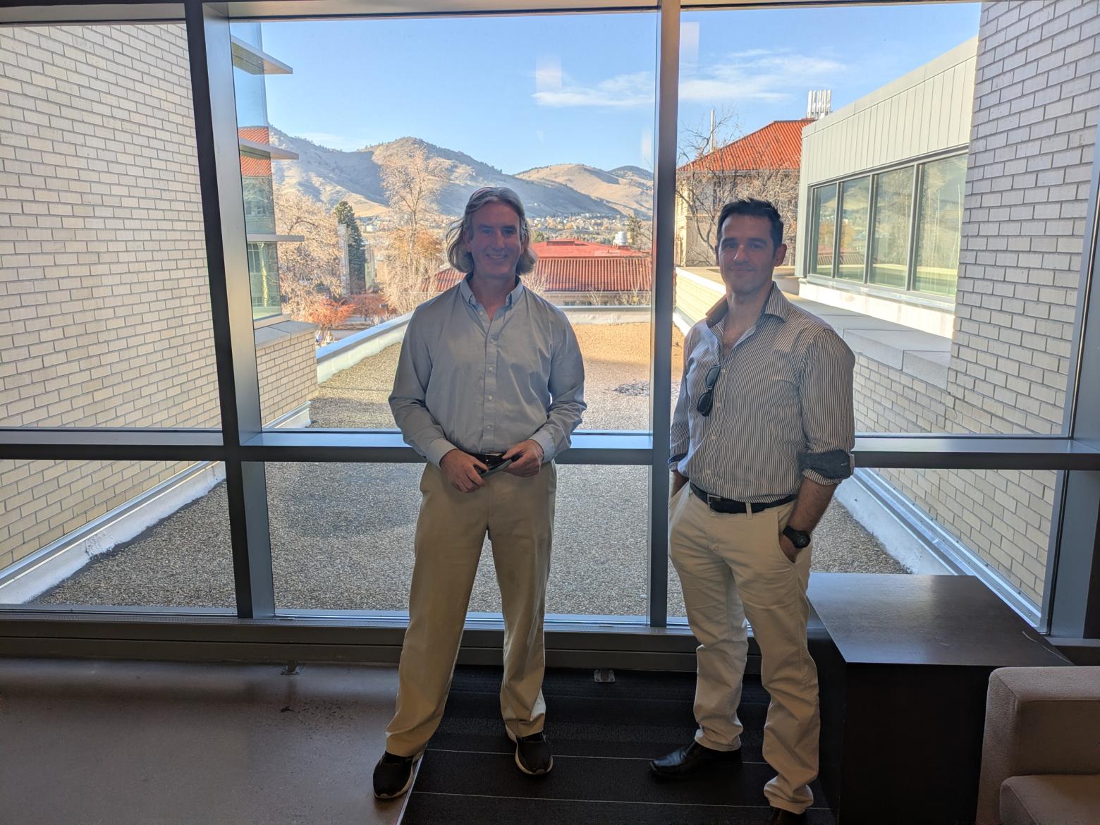 Andy Osborne and Mark Deinert stand in front of a window with campus buildings in the background