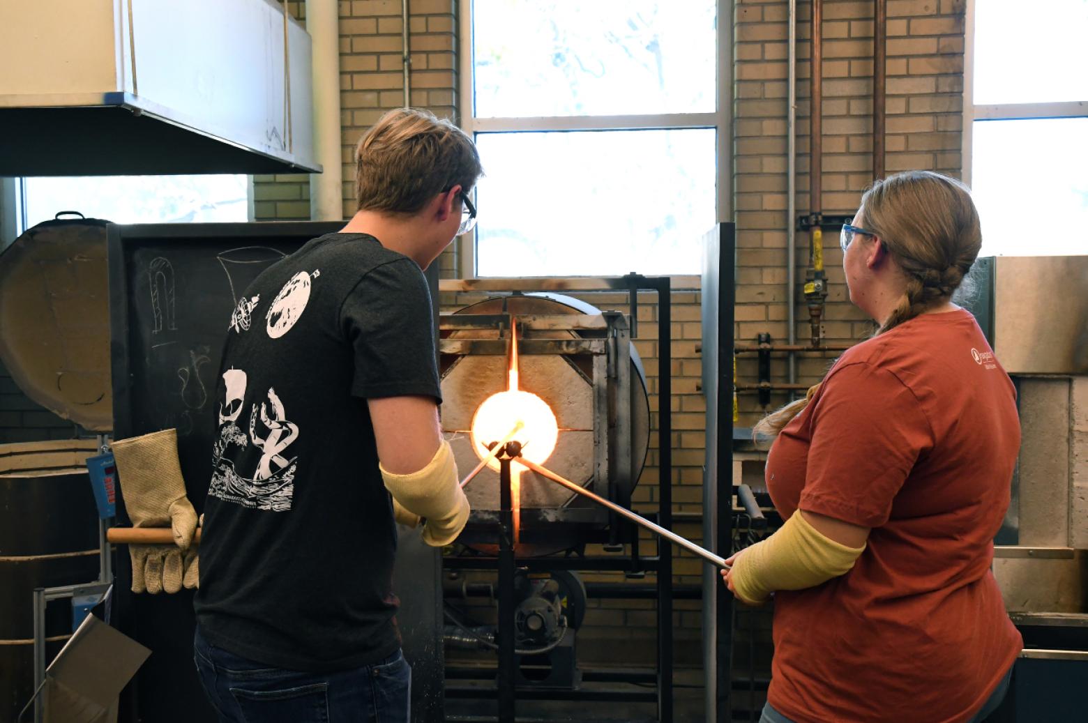 Two Mines students wearing protective gloves shaping molten glass in a furnace-lit glassblowing workshop.