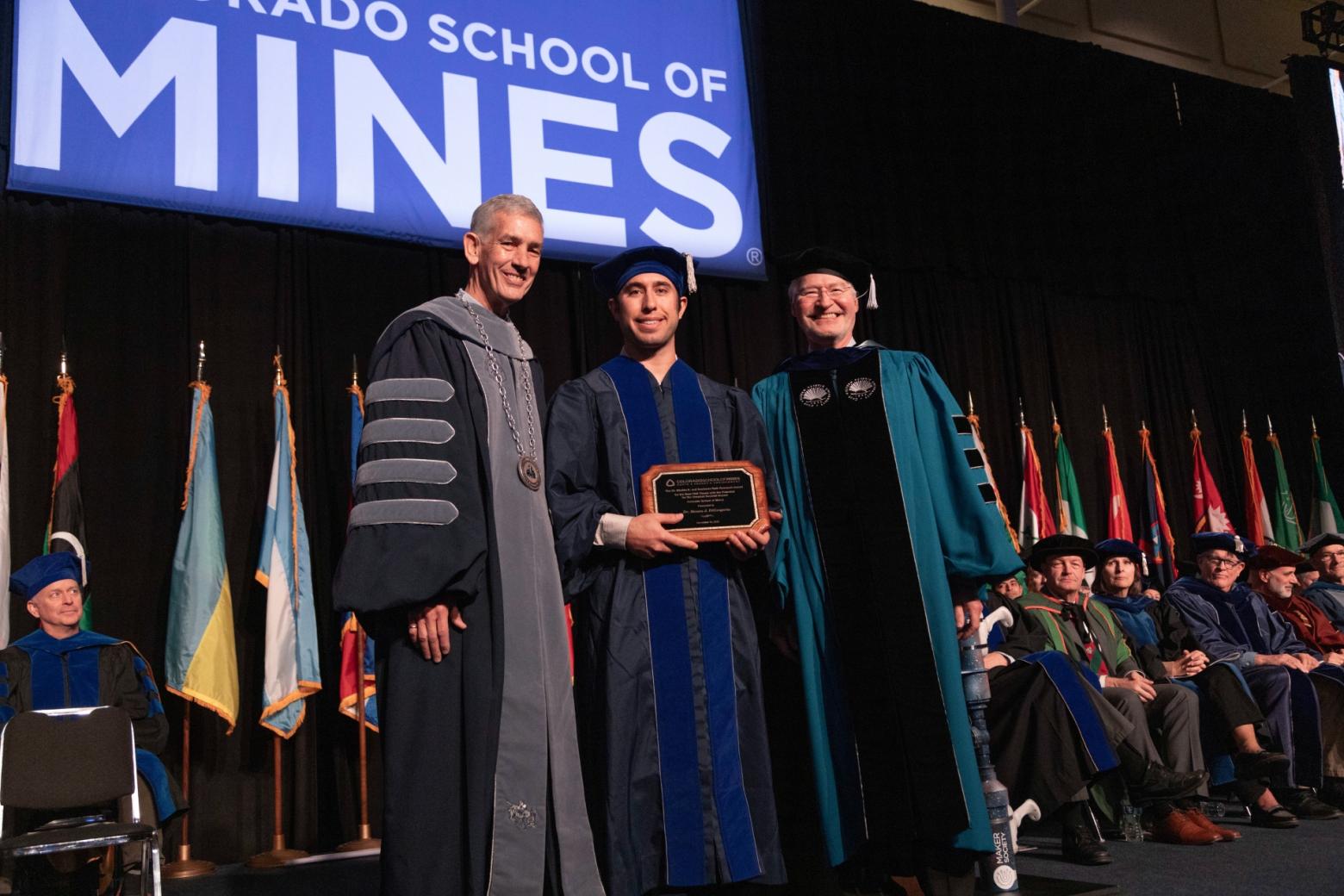 Steven DiGregorio stands on stage at the 2023 Mines commencement in graduation robes with President Paul C Johnson
