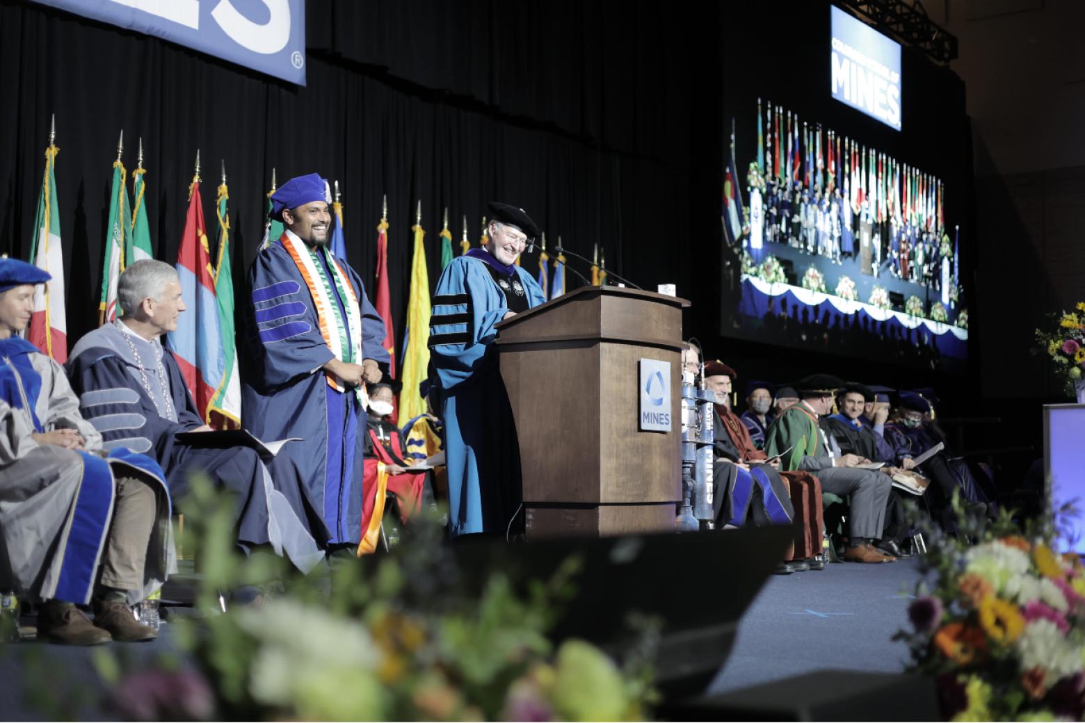 A PhD graduate stands next to a podium where the VP of research is speaking
