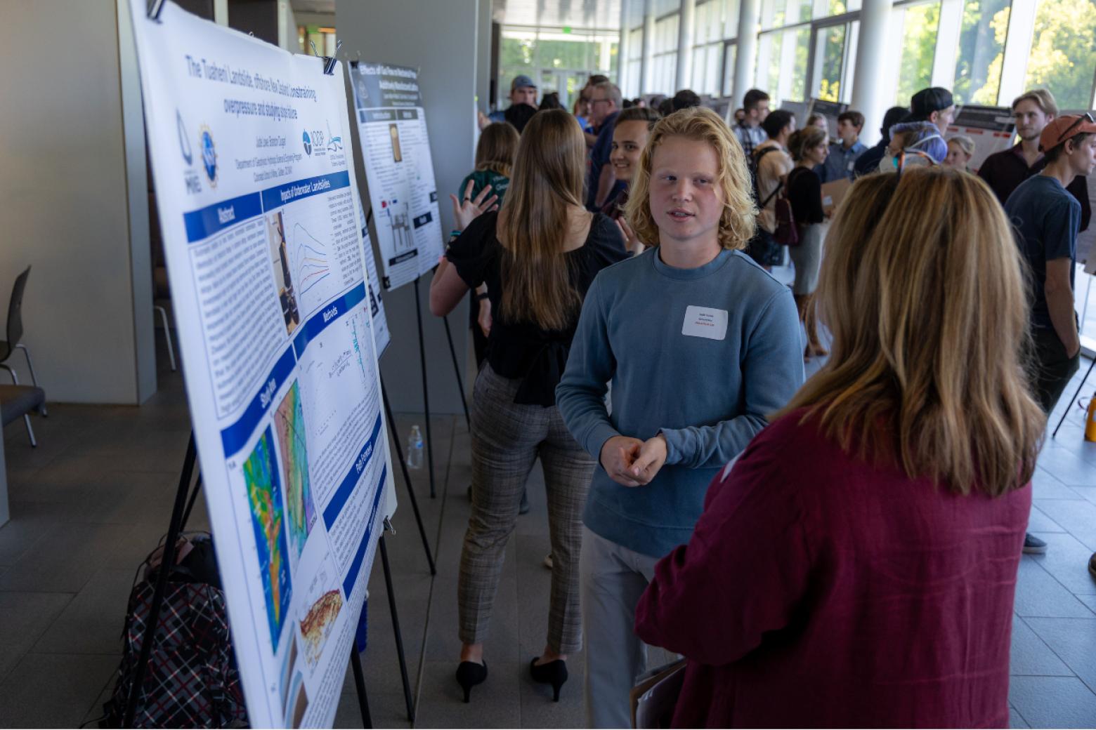 A Mines undergraduate student presents his research, referring to a poster board on an easel, to an onlooker.