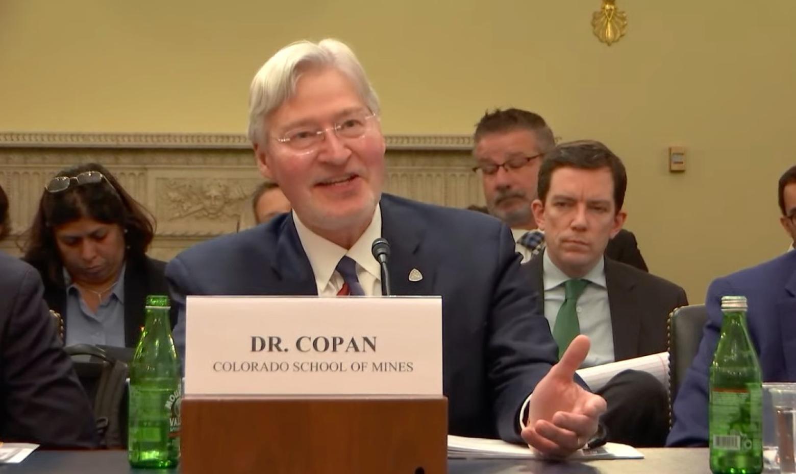 Walt Copan sits at a table behind a sign with his name on it speaking