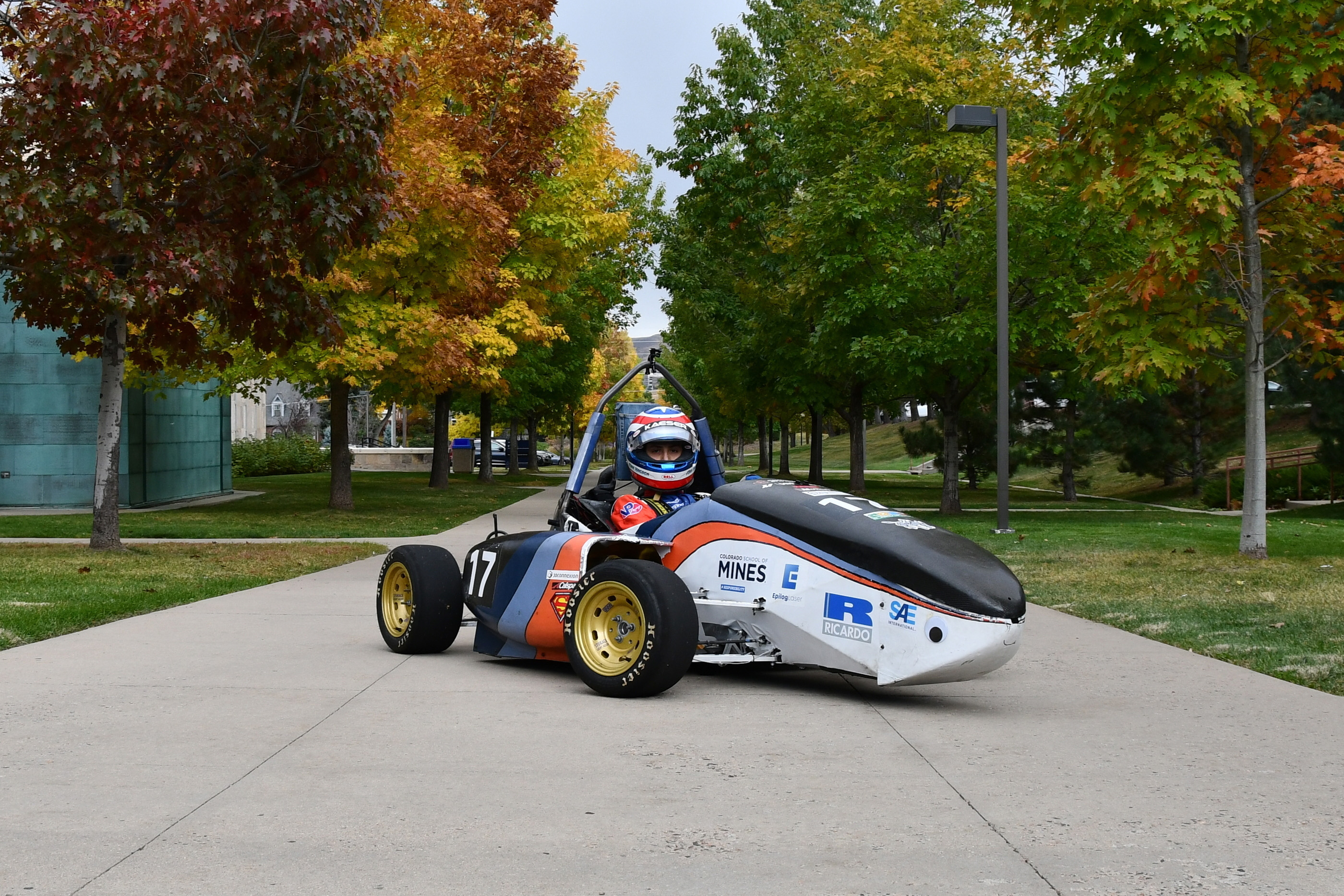 Formula SAE race car parked on a campus walkway, with a driver seated in the open cockpit. The car is numbered 17 and bears Colorado School of Mines branding. Trees with fall foliage surround the walkway.