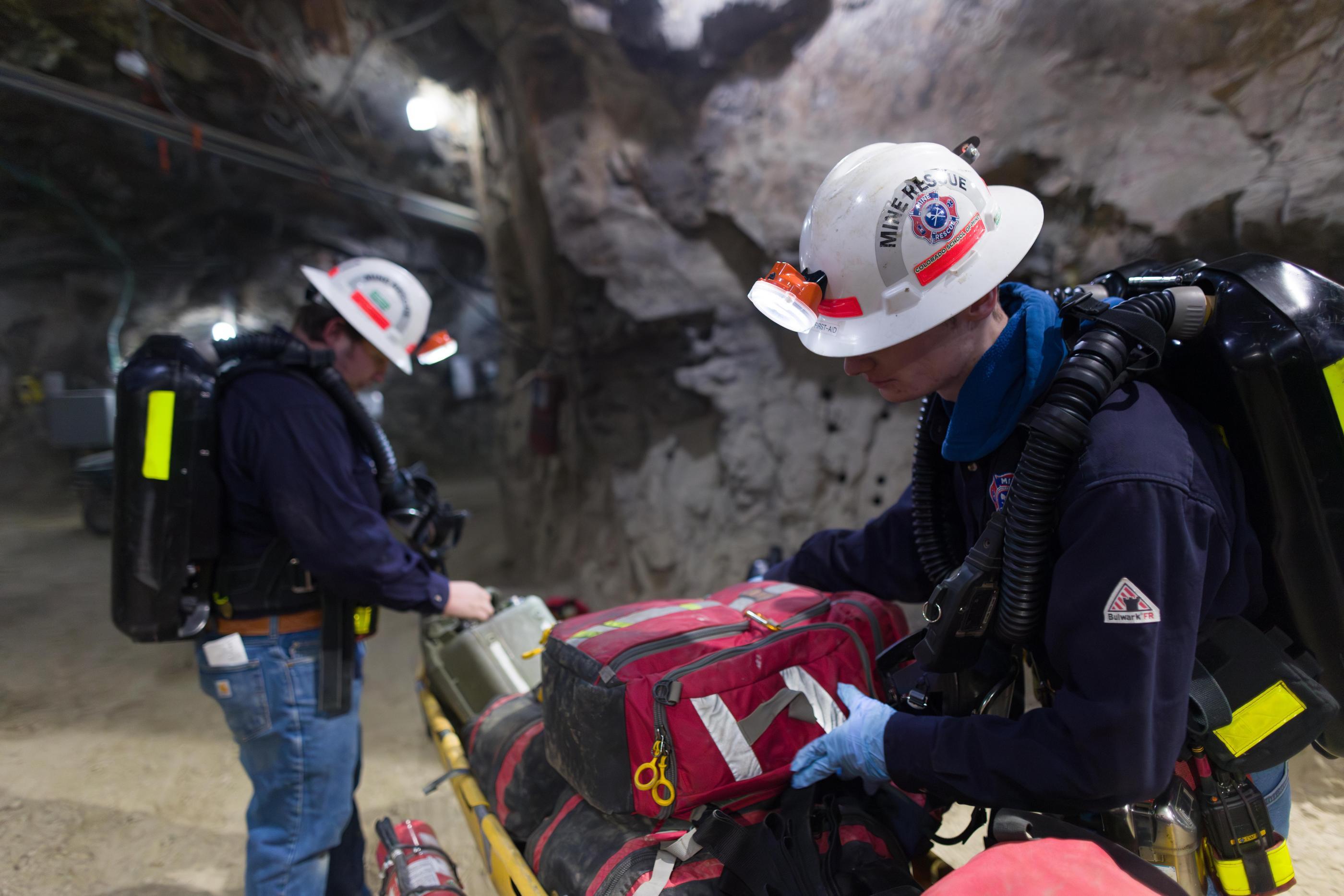 Two mine rescue team members in helmets and breathing apparatus prepare emergency equipment inside an underground mine.