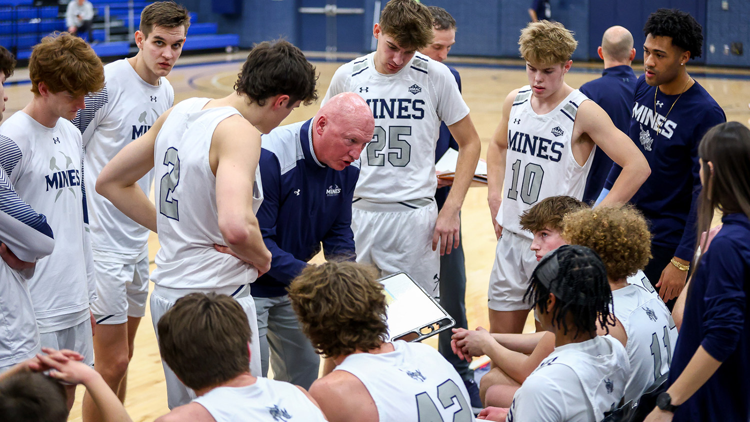 Men's basketball coach Pryor Orser holds a clipboard and stands in a huddle of basketball players