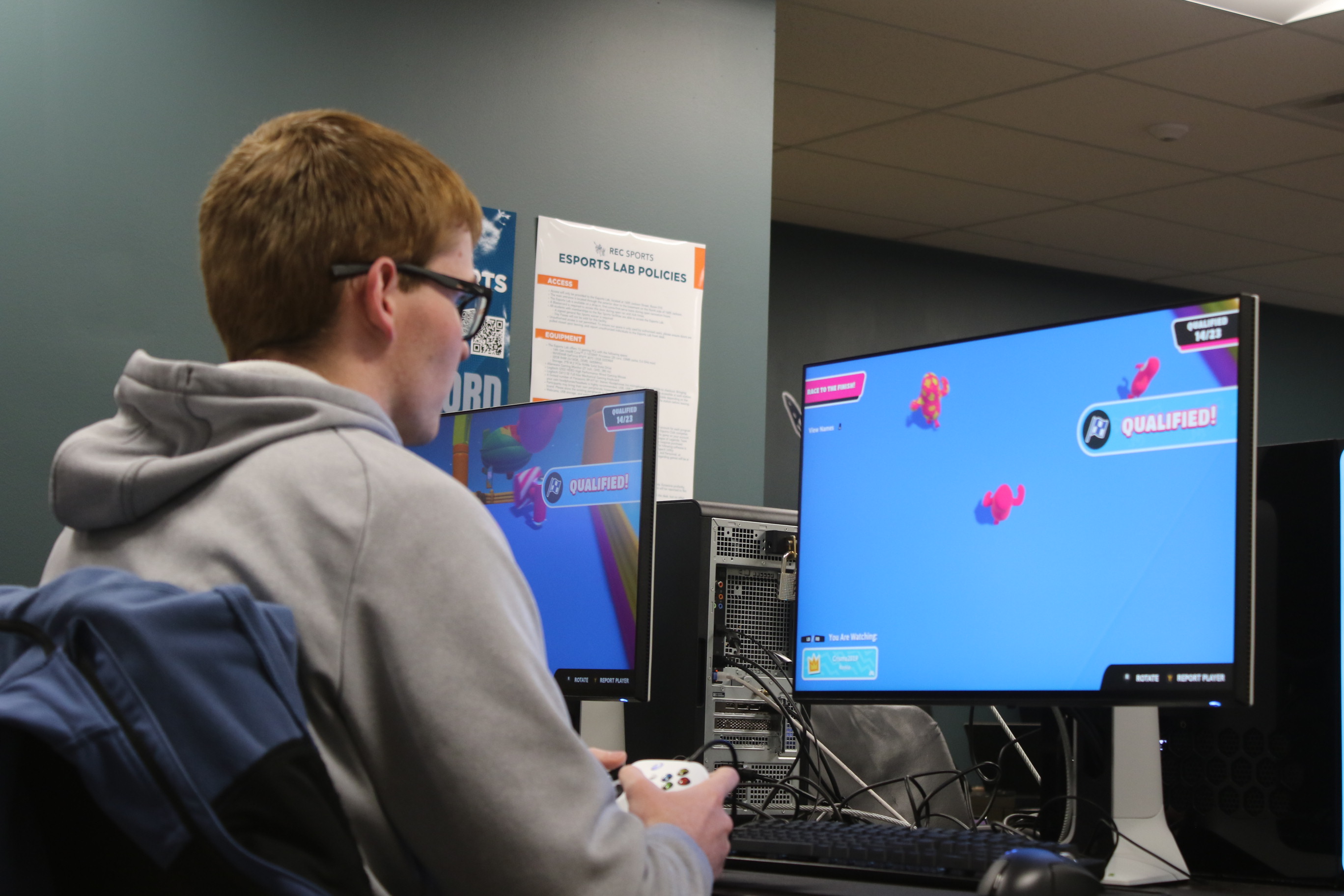 A student in a gray hoodie and holding a gaming controller sits in front of a computer screen