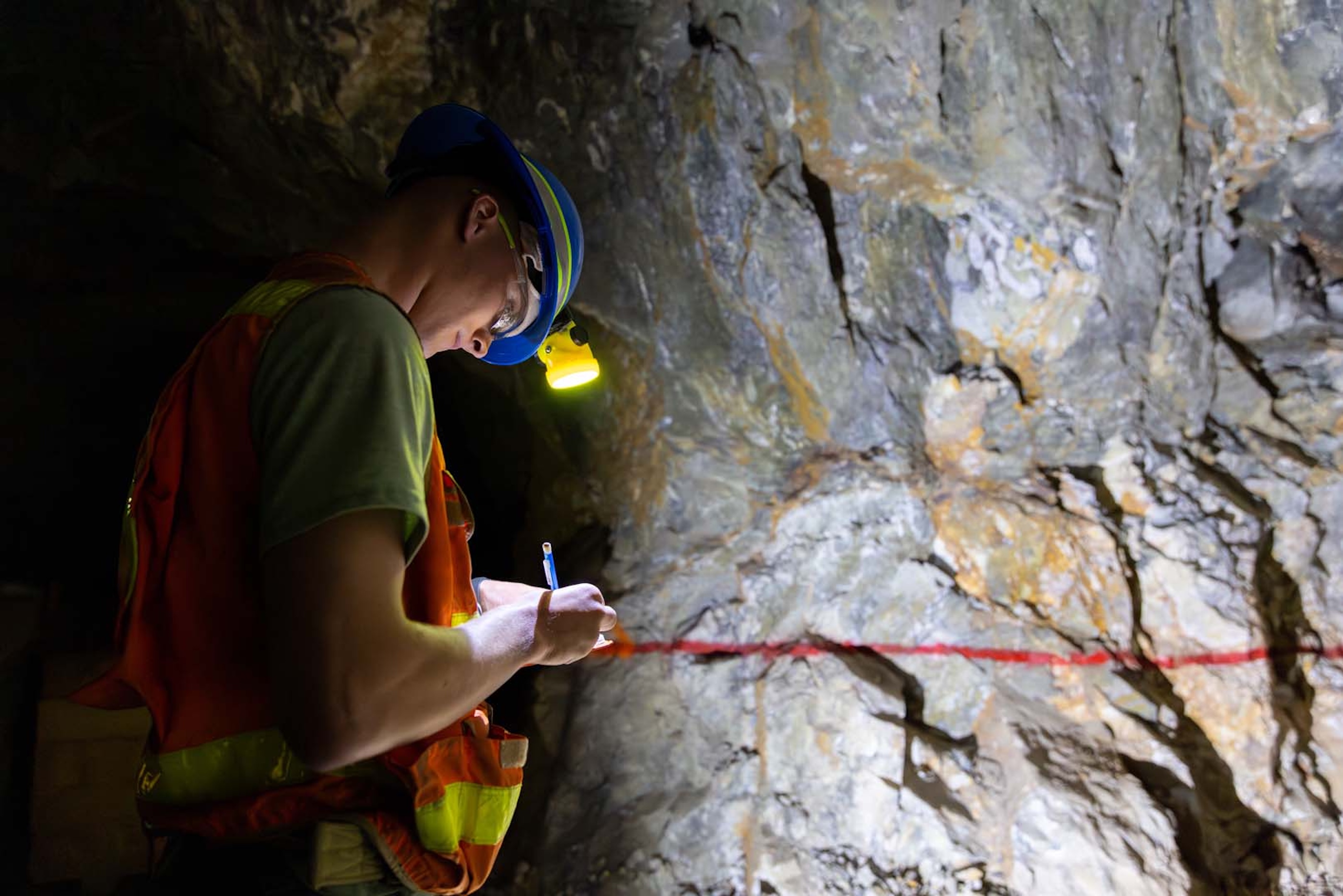 A student wearing a hard hat and safety vest writes in a notebook in front of a rock face within Edgar Mine.