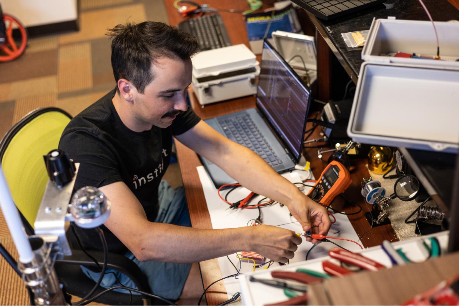 Trevor Bachand sits at a desk with a laptop and works with a device with wires