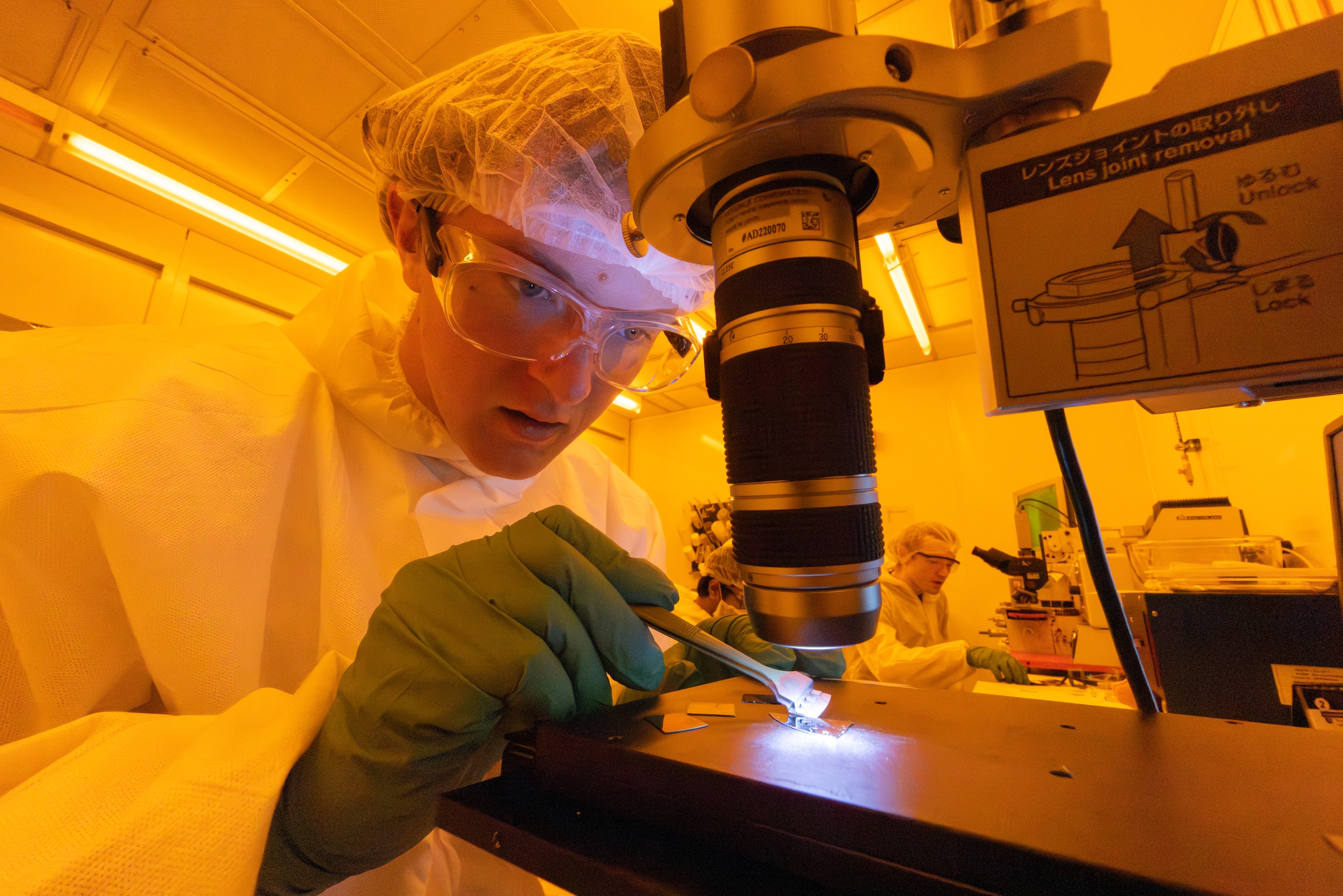A student in a bunny suit and safety goggles inspecting samples within a lab tinted with yellow lighting.