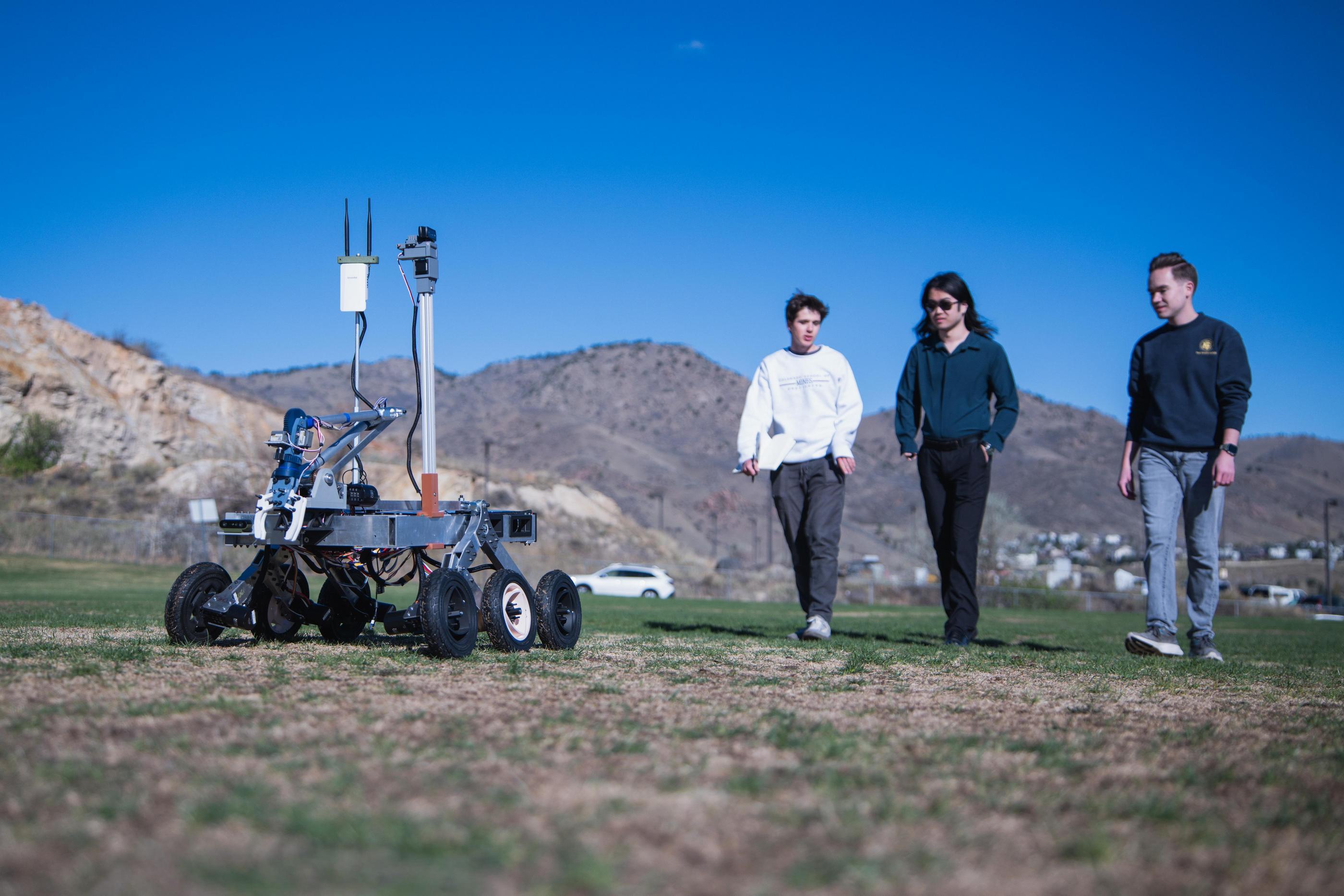 Students walk alongside a six-wheeled rover equipped with sensors and cameras on a grassy field, with mountains in the background.