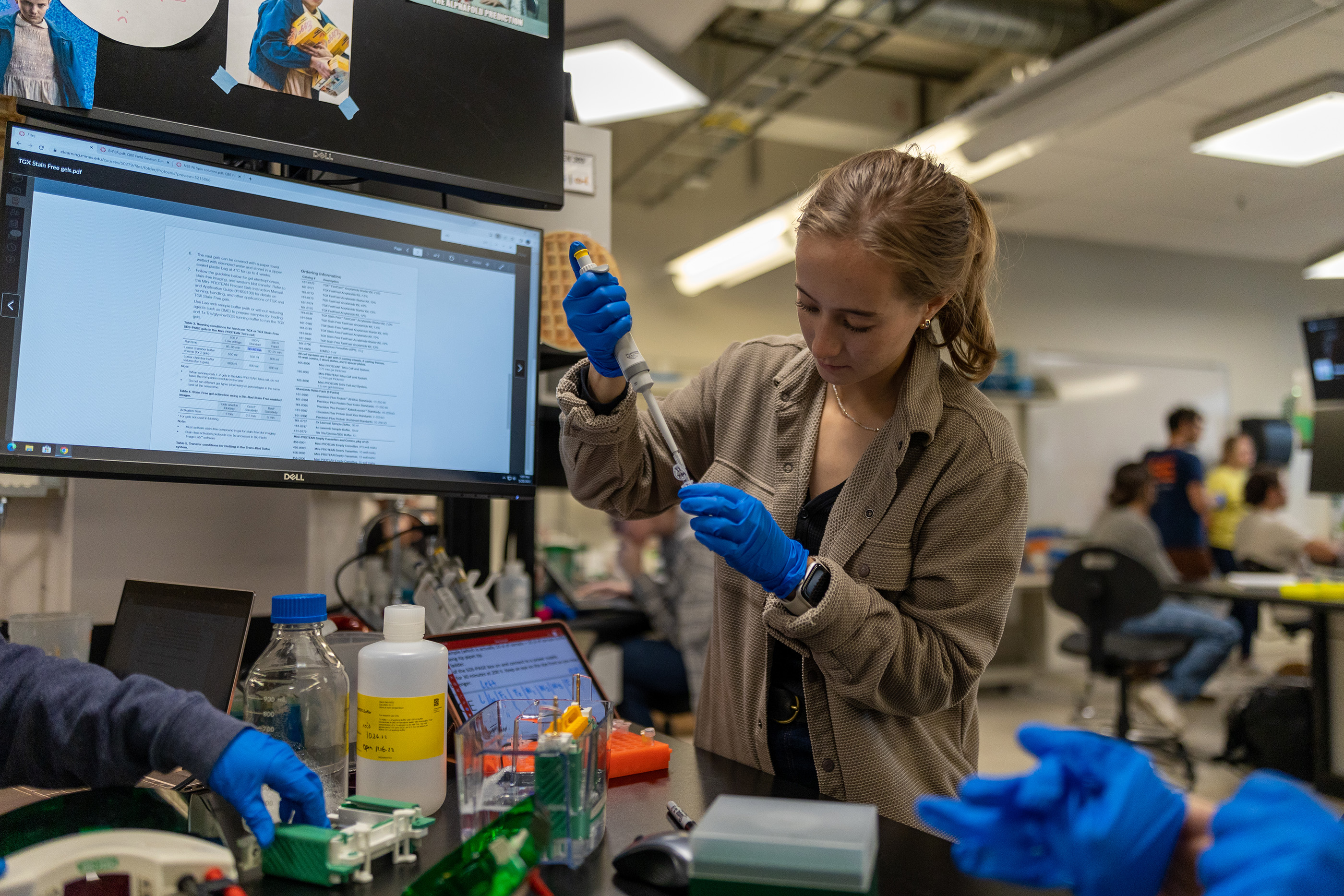 A female student wearing blue gloves holds a pipette in a bioscience lab