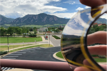 A hand bends a sample of a dynamic tinted window showing the reflection of a parking lot