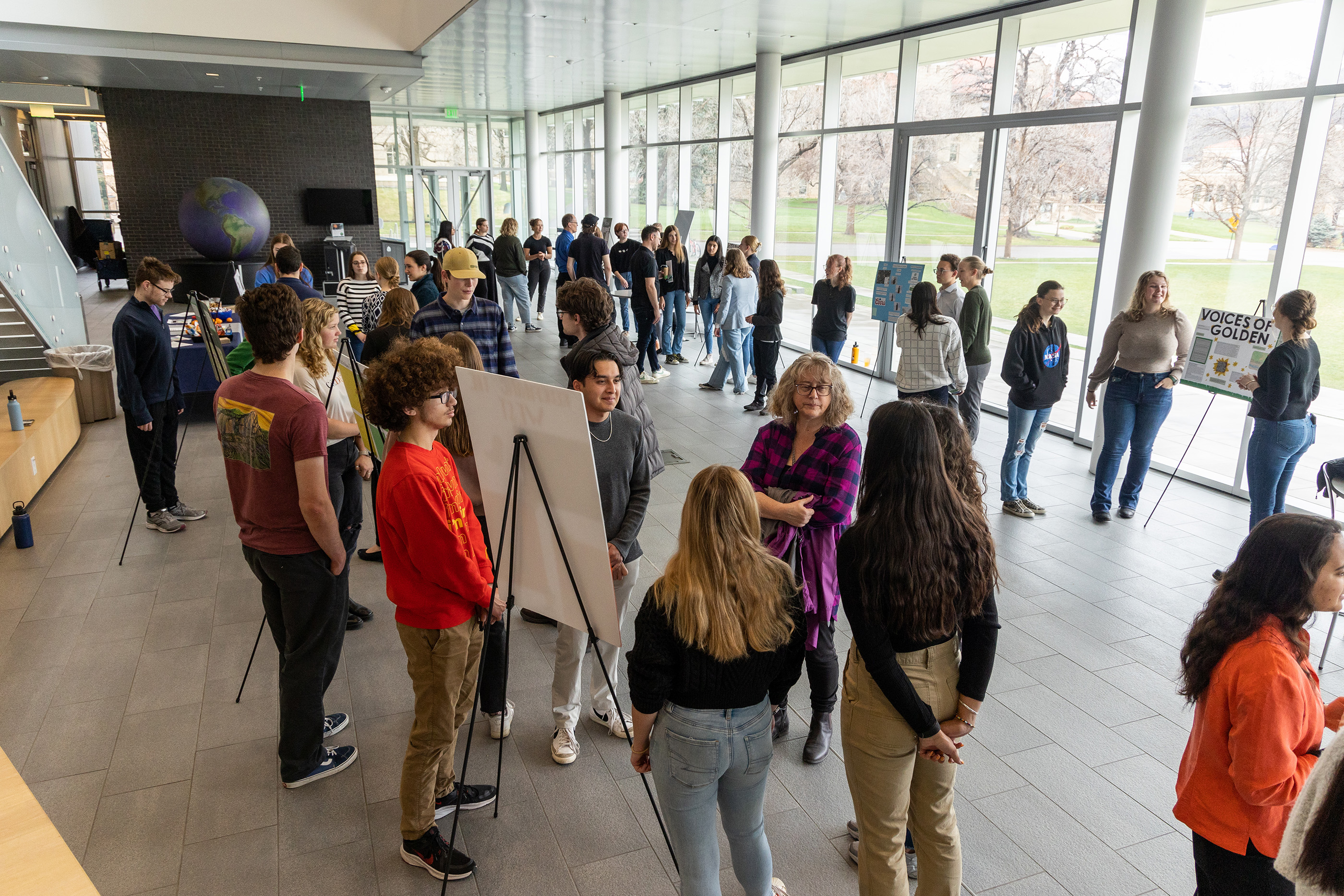 Students stand near posters in the CoorsTek atrium presenting projects to event attendees