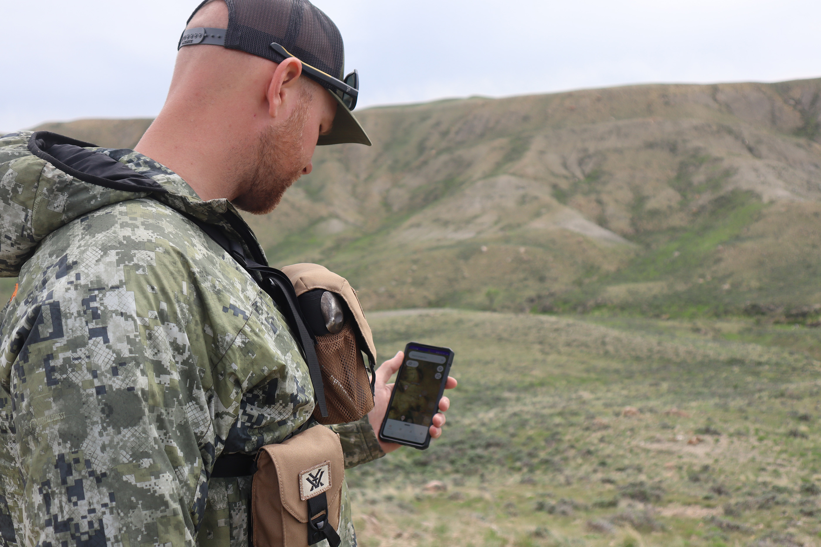 Man in a camo jacket stands outdoors holding a phone showing a map interface