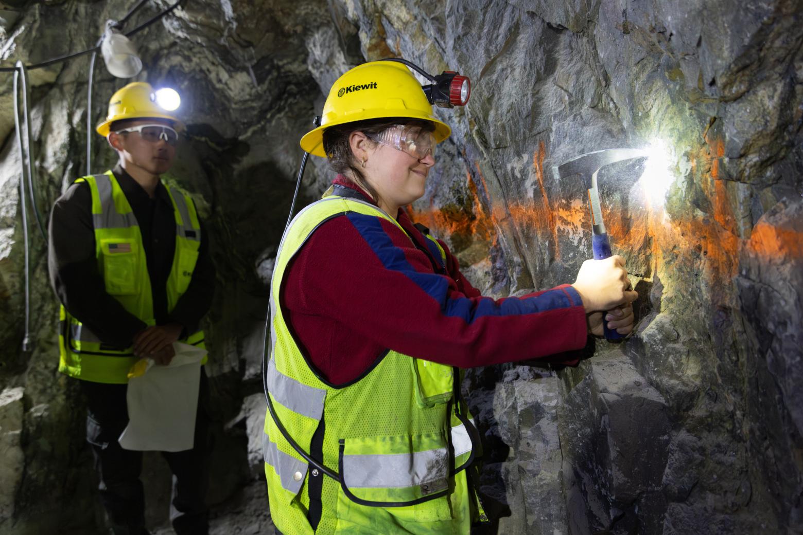 A student in a hard hat, safety vest and safety goggles chisels a rock face in Edgar Mine with another student dressed similarly standing behind