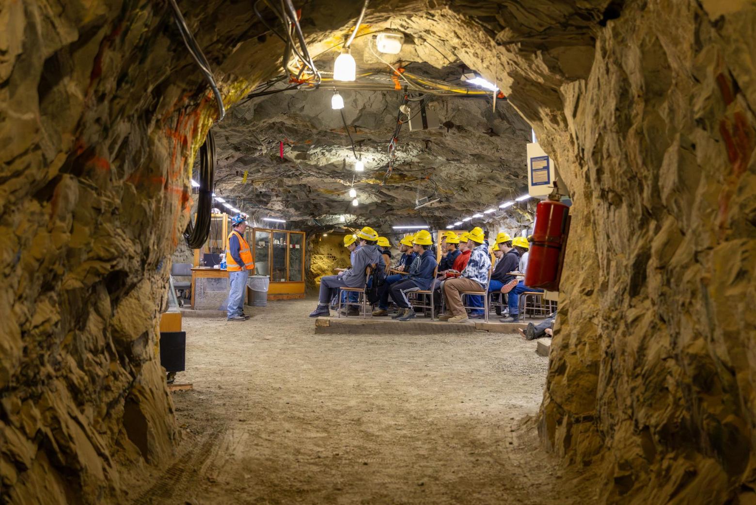 An underground classroom with students in hard hats at desks listening to a professor.
