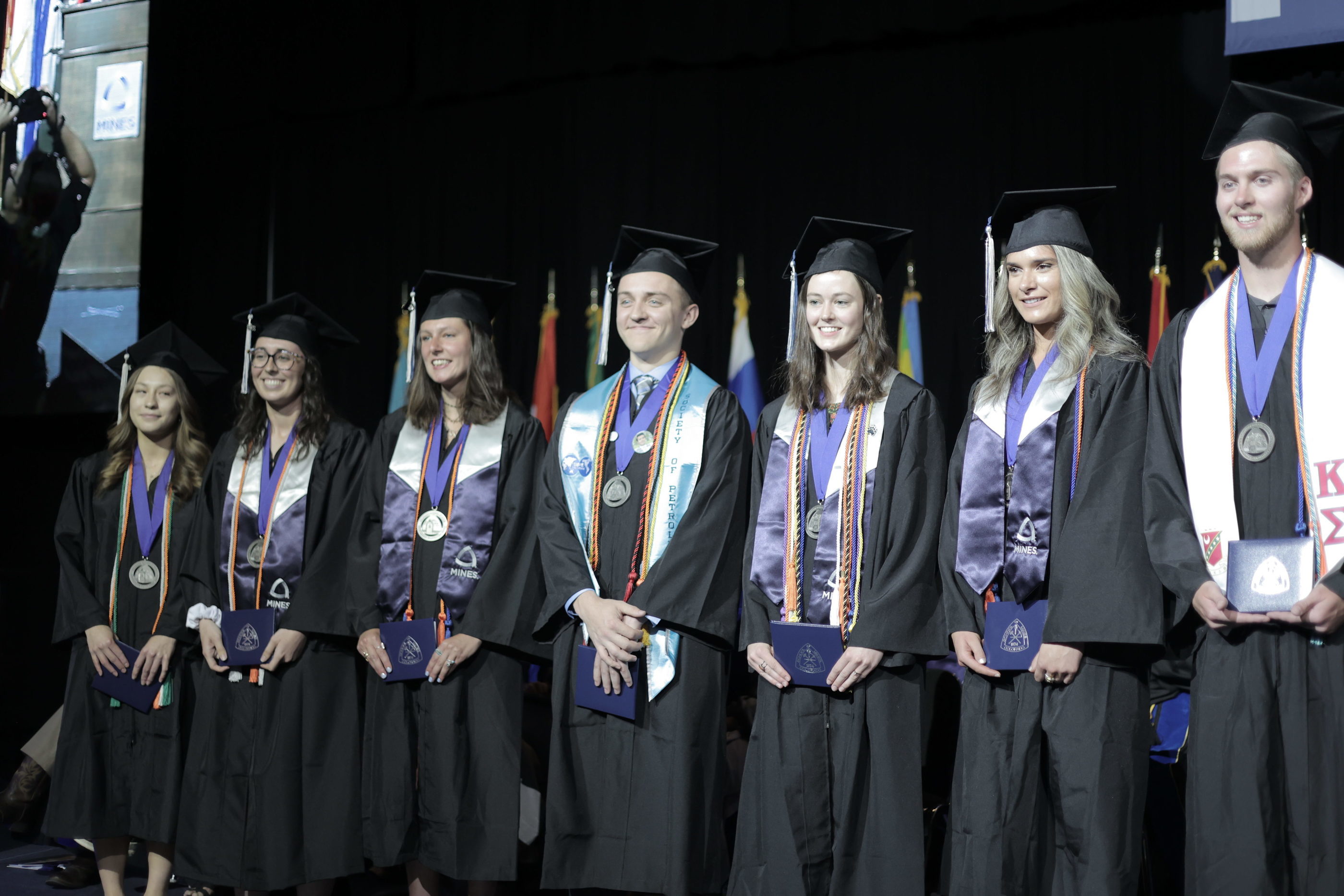 A group of college graduates wearing a medal and graduation cords around their necks stands in a line